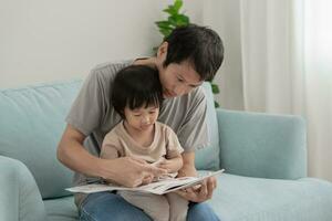 contento asiático padre relajarse y leer libro con bebé hora juntos a hogar. padre sentar en sofá con hija y leyendo un historia. aprender desarrollo, cuidado de niños, reír, educación, contar historias, práctica. foto