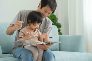 Happy Asian father relax and read book with baby time together at home. parent sit on sofa with daughter and reading a story. learn development, childcare, laughing, education, storytelling, practice. photo