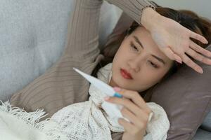 Young Asian woman having high fever while checking body temperature, female sneezing and runny nose with seasonal influenza, allergic, digital thermometer, virus, coronavirus, illness, respiratory photo