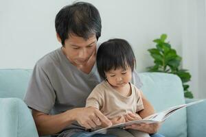 contento asiático padre relajarse y leer libro con bebé hora juntos a hogar. padre sentar en sofá con hija y leyendo un historia. aprender desarrollo, cuidado de niños, reír, educación, contar historias, práctica. foto