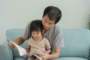 contento asiático padre relajarse y leer libro con bebé hora juntos a hogar. padre sentar en sofá con hija y leyendo un historia. aprender desarrollo, cuidado de niños, reír, educación, contar historias, práctica. foto