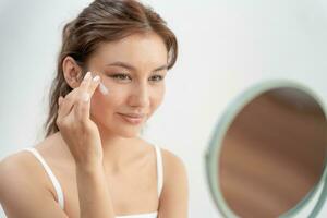 Beautiful Asian woman sit in front of a mirror. face of a healthy woman applying cream and makeup. Advertisement for skin cream, anti-wrinkle, baby face, whitening, moisturizer, tighten pores serum photo