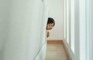 pequeño asiático niños niño , jugando esconder y buscar en pie siguiente un pared casa, adorable niño teniendo divertido en el hogar. contento familia tener divertido con niños, actividad, aprendizaje foto