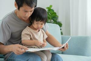 contento asiático padre relajarse y leer libro con bebé hora juntos a hogar. padre sentar en sofá con hija y leyendo un historia. aprender desarrollo, cuidado de niños, reír, educación, contar historias, práctica. foto