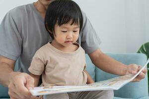 padre relajarse y leer libro con bebé hora juntos a hogar. padre sentar en sofá con hija y leyendo un historia. aprender desarrollo, cuidado de niños, reír, educación, contar historias, práctica. foto