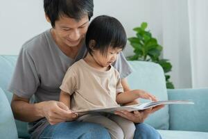 Happy Asian father relax and read book with baby time together at home. parent sit on sofa with daughter and reading a story. learn development, childcare, laughing, education, storytelling, practice. photo