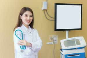 Professional young Asian woman doctor wearing white robe and stethoscope standing with arms crossed happy and smile at examination room in hospital. photo