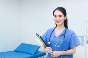 Professional young Asian woman doctor standing smiling in medical uniform blue lab shirt holding patient documents in hand examination room in the hospital. Health care concep. photo