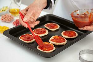 Senior woman adding pizza sauce to the stretched dough. Delicious homemade mini pizzas preparation. photo