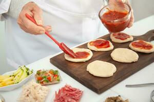 Senior woman adding pizza sauce to the stretched dough. Delicious homemade mini pizzas preparation. photo