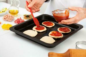 Senior woman adding pizza sauce to the stretched dough. Delicious homemade mini pizzas preparation. photo