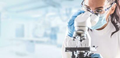 Female lab technician in protective glasses, gloves and face mask sits next to a microscope in laboratory photo