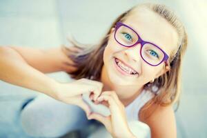 Smiling little girl with dental braces and glasses showing heart with hands photo