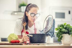 Woman holding spoon and lid is cooking at home, opening saucepan that fogs her glasses photo