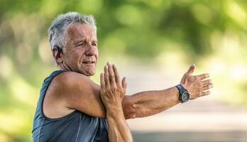 Elderly fit man stretches his arm on a summer day outdoors, wearing watches and earphones photo