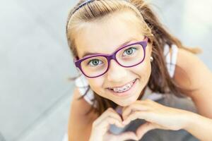 joven preadolescente niña en lentes vistiendo tirantes sonrisas a el cámara demostración corazón forma con su manos foto