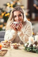 retrato de un joven rubia sentado a un Navidad mesa en frente de un Navidad árbol, Bebiendo caliente café, té o puñetazo foto