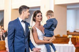 Happy young newlyweds stand in the church with the bride holding a young son in her arms photo