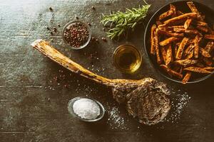 Grilled beef tomahawk steak with sweet potato fries salt spices olive oil and rosemary on rustic board - top of view photo