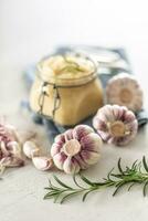 Homegrown garluc bulbs in front of a glass jar full of fresh garlic paste laid on kitchen cloth with rosemary photo