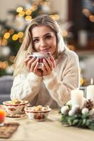 retrato de un joven rubia sentado a un Navidad mesa en frente de un Navidad árbol, Bebiendo caliente café, té o puñetazo foto