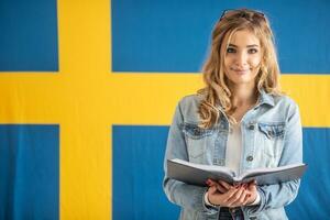 Teenage student with open book stands with Swedish flag behind her photo