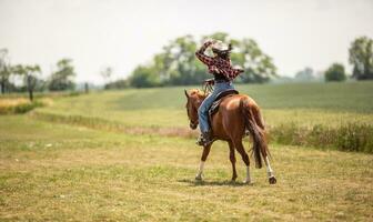 Woman in jeans shirt holds her cowgirl hat as she gallops on a meadow on her paint horse photo