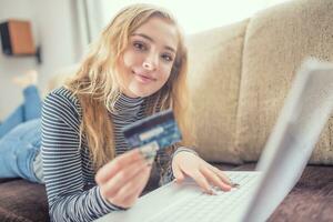 Beautiful young girl holding credit card buying online at sofa in living room photo