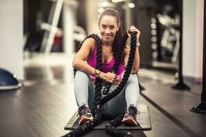 Woman holding battle ropes sits on a mat nside the gym, smiling at the camera photo