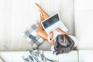 parte superior ver de hogar oficina mujer, trabajando en un computadora portátil, inclinado en contra un blanco sofá, mirando dentro el ordenador personal, desnudo piernas, su célula teléfono siguiente a su en un gris y blanco comprobado almohada foto