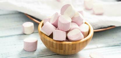 Close up of a wooden bowl full of pink and white marshmallows with some scattered around on a white table cloth, dark tray and white wooden table photo