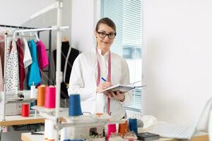 Young female business owner takes notes into her notepad while standing in her tailor workshop photo