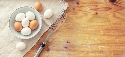 Rustic cutting board and tablecloth with nine eggs in and out of a bown and a metal steel whisk photo