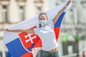 Slovak female fan holds a flag behind her on a street, wearing a face mask photo