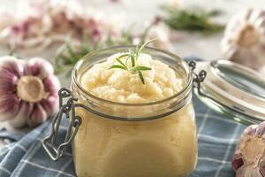 Aromatic garlic paste in a glass jar laid on rustic kitchen cloth with bulbs and peeled cloves and rosemary photo