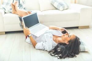 Dark-haired good looking girl lying on her back on the floor with her feet up on a white sofa, a pillow under her head, holding a laptop and having a phone call photo