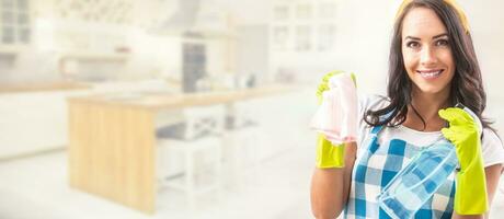 Banner view of a smiling beautiful woman in apron holding detergent and cleaning rag in her hands in rubber gloves in the right third of the picture with kitchen in the background photo