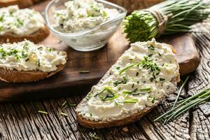 Slices of crusty bread and glass bown with a cream cheese spread and freshly cut chives on a vintage wooden cutting board photo