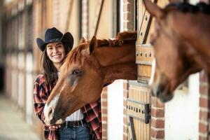 dos caballos y un joven mujer en vaquero sombrero siguiente a cada otro en el establos foto