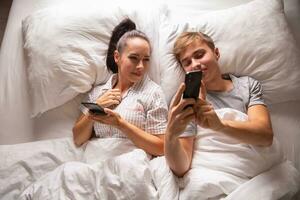 Top view of a couple sharing interesting and funny things on their mobile phones while lying in white bedding before sleep photo