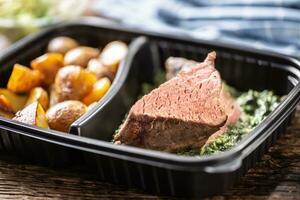 Delicious red meat sous vide steak packed in portable take away plastic box and served with crunchy roasted potatoes and savory spinach dip and fresh vegetable salad photo