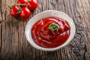 Ketchup or tomato sauce in white bowl and cherry tomatoes on wooden table. photo