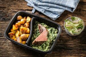 Delicious red meat sous vide steak packed in portable take away plastic box and served with crunchy roasted potatoes and savory spinach dip and fresh vegetable salad photo