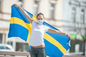 Beautiful smiling Swedish girl holds a flag while wearing a protective face mask outdoors photo