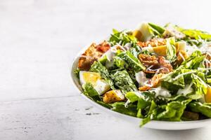 Caesar salad prepared from romaine lettuce, baked bacon, egg, bread croutons, garlic dressing and grated Parmesan cheese on white table. photo