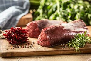 Raw pieces of red sirloin meat on a wooden board next to fresh rosemary and chilli peppers photo