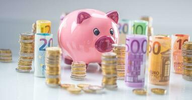 Pink piggy bank in the middle of rolled euro banknotes and towers with coins photo