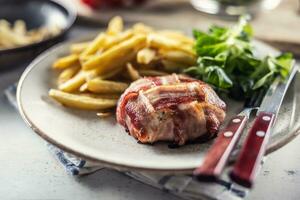 Bacon wrapped meat served with french fries and salad on a plate with cutlery photo