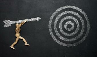 A wooden mannequin holds a drawn arrow and aims precisely at the target on a black chalkboard. The concept of achieving set goals photo