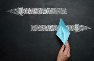 A woman's hand holds a blue paper boat on a black chalk board and arrows in different directions, the choice of the direction of movement photo